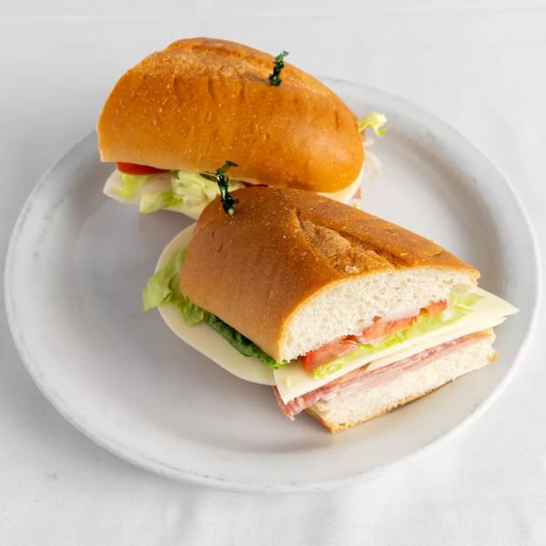 Two deli sandwiches on baguettes with meat, cheese, lettuce, and tomato on a white plate.