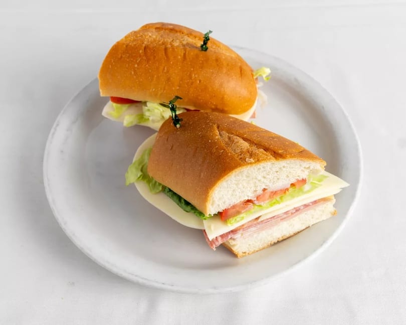 Two deli sandwiches on baguettes with meat, cheese, lettuce, and tomato on a white plate.