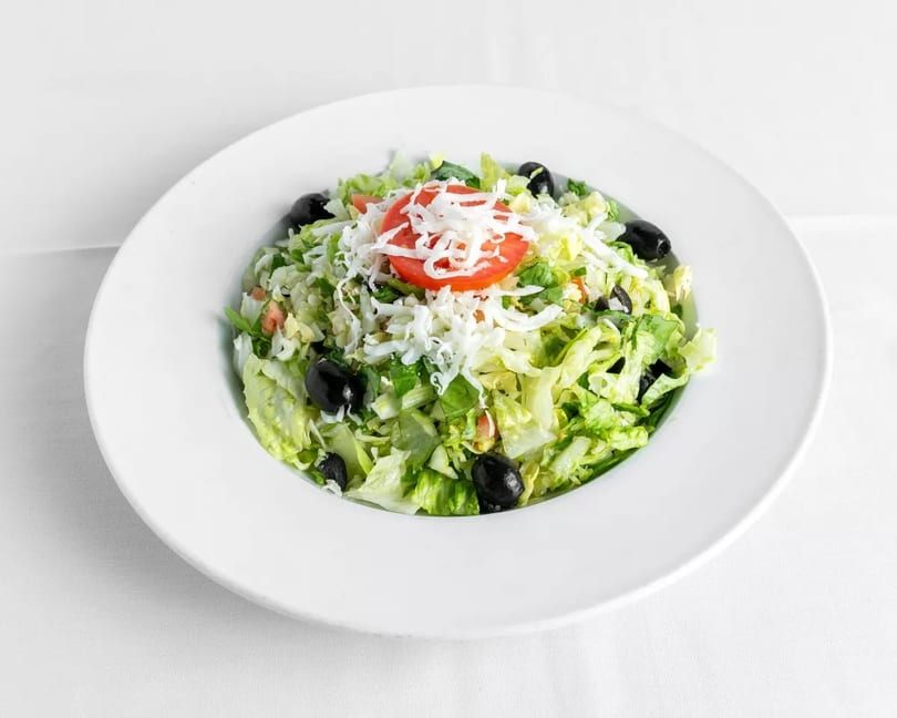 A fresh garden salad with lettuce, black olives, shredded cheese, and a tomato slice in a white bowl.