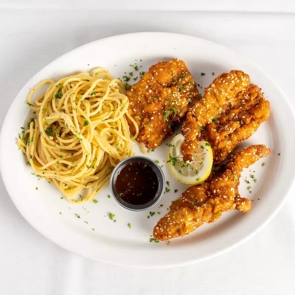 Crispy sesame chicken tenders with spaghetti, a lemon slice, and dipping sauce on a white plate.