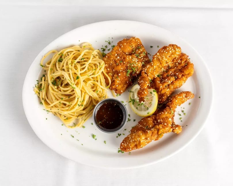 Crispy sesame chicken tenders with spaghetti, a lemon slice, and dipping sauce on a white plate.