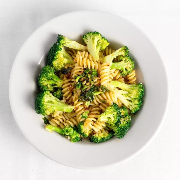 A white bowl filled with fusilli pasta, broccoli florets, and green herbs on a white tablecloth.