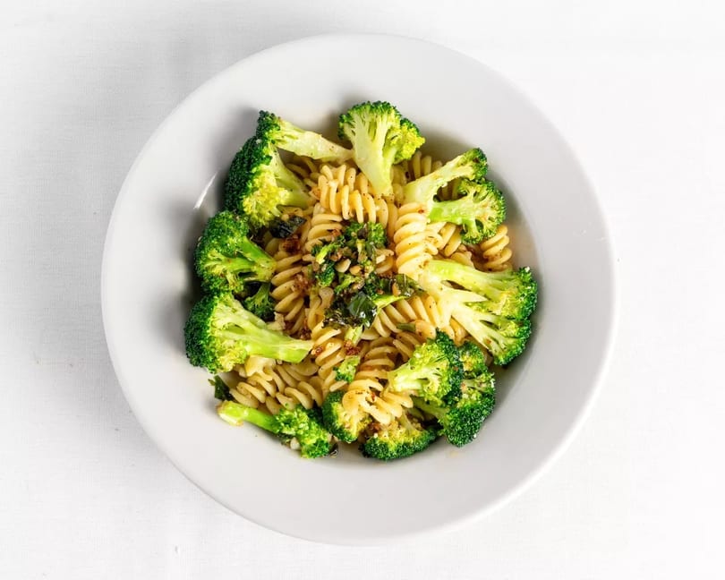 A white bowl filled with fusilli pasta, broccoli florets, and green herbs on a white tablecloth.