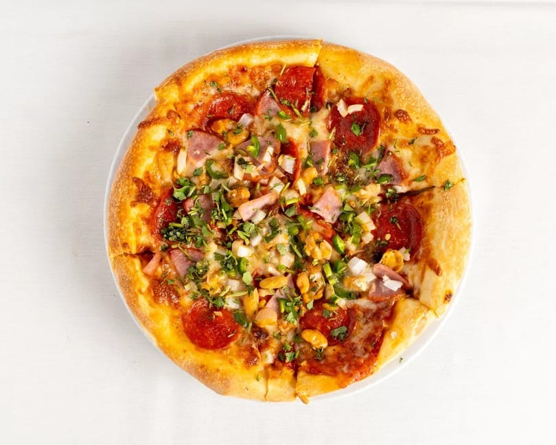 A whole pizza with pepperoni, ham, onions, green peppers, and cilantro on a white plate.