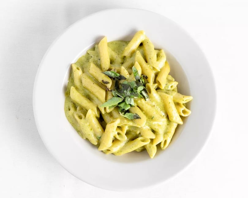 Penne pasta with creamy green pesto sauce and fresh basil in a white bowl.