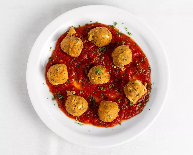 Seven golden-brown meatballs in a rich red tomato sauce, garnished with fresh parsley on a white plate.